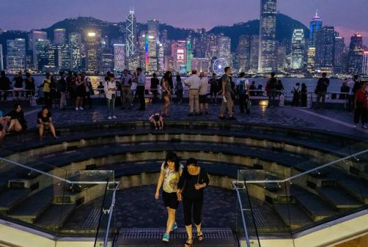 In this undated photo, people look out from the waterfront in the Tsim Sha Tsui district as buildings across the Victoria Harbour stand illuminated at night in Hong Kong. (ANTHONY KWAN / BLOOMBERG)