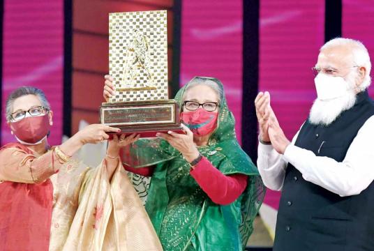 Prime Minister Sheikh Hasina and her younger sister Sheikh Rehana hold high the Gandhi Peace Prize-2020 awarded to Bangabandhu Sheikh Mujibur Rahman posthumously by the Indian government. Indian Prime Minister Narendra Modi is seen applauding next to them on the final day of twin celebrations at the National Parade Square yesterday. Photo: PID